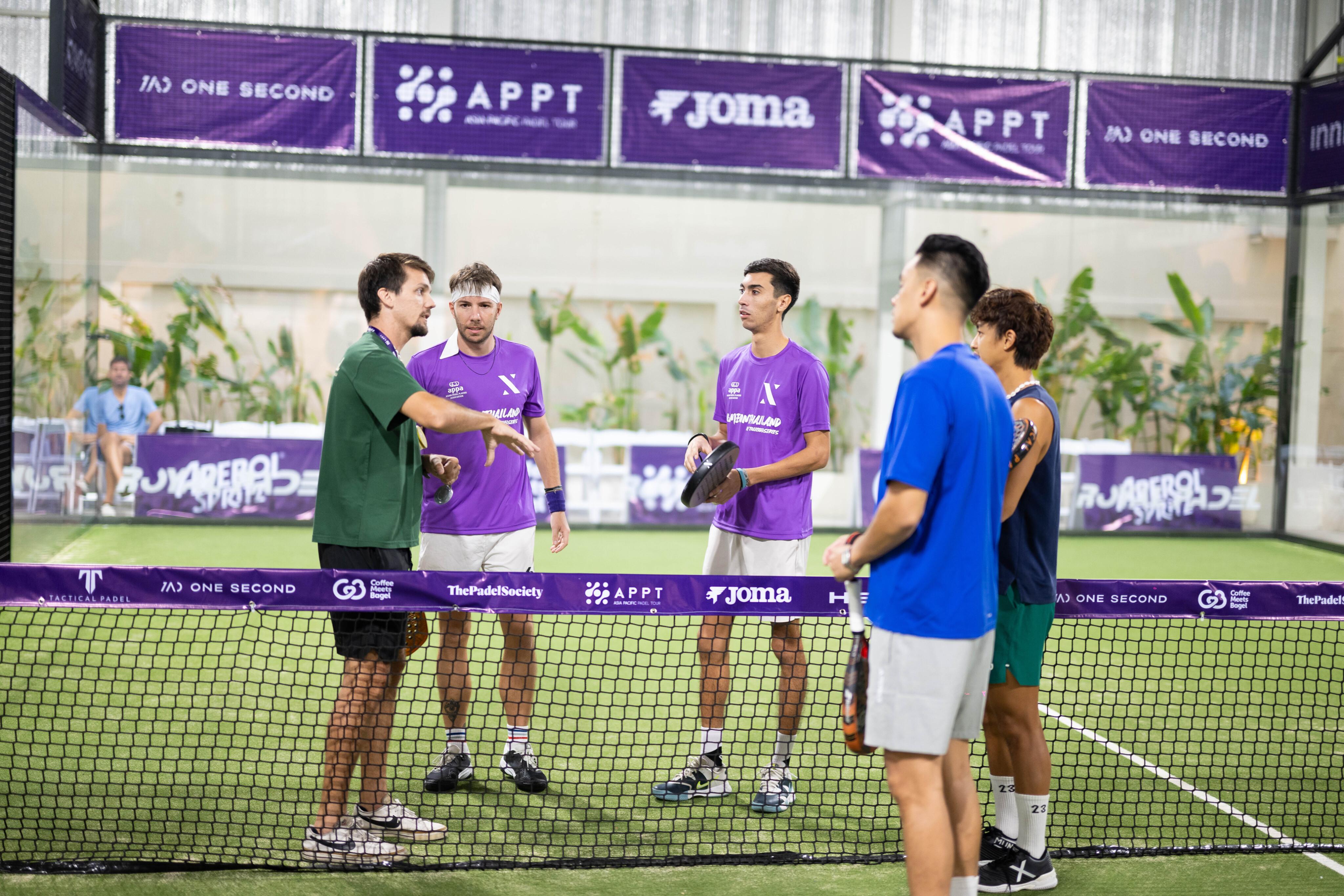 A group of four padel players standing by the net on an indoor padel court, listening to a padel coach in a green shirt