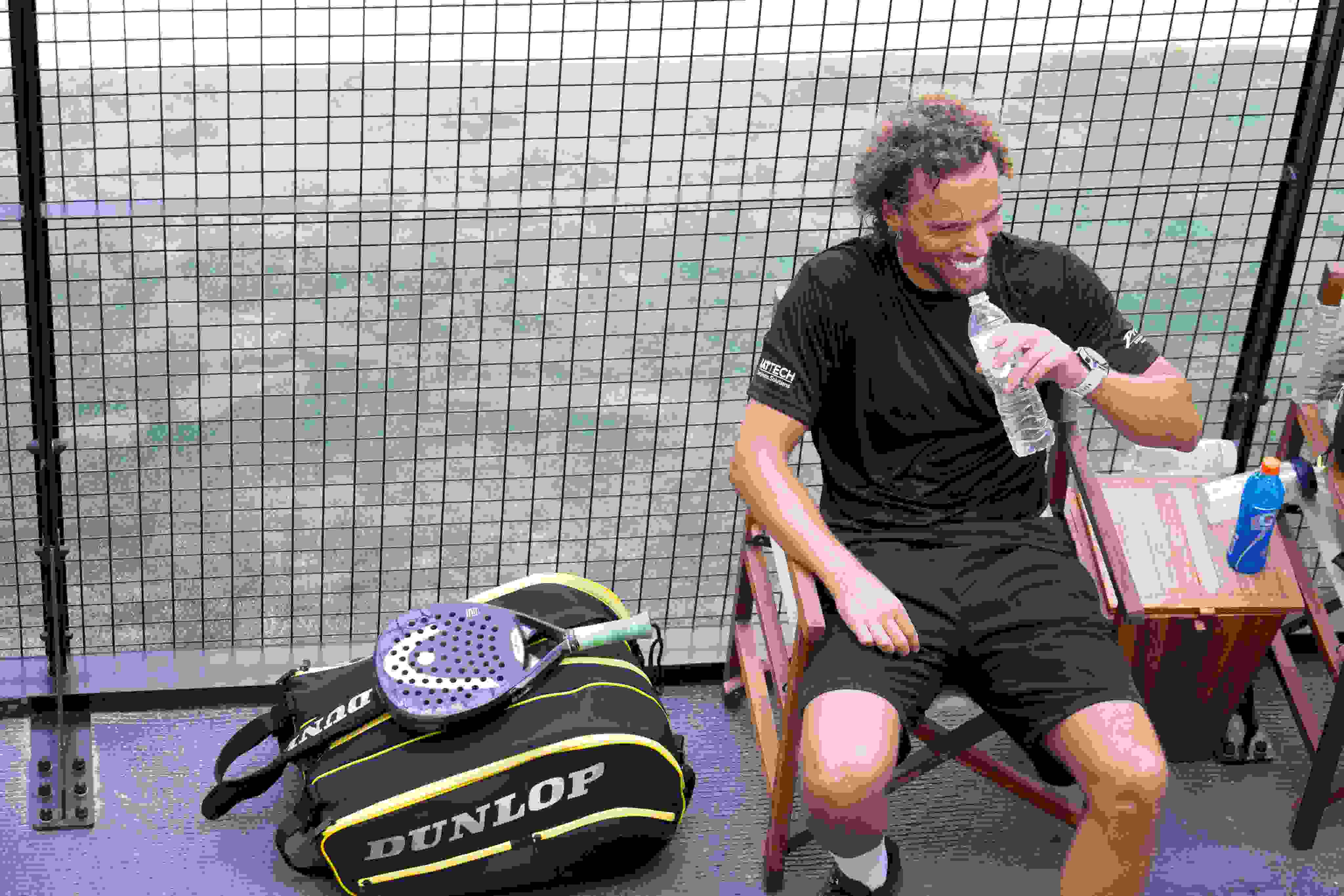 A padel player in a black shirt sits on a wooden chair, smiling while drinking water with a sport bag and padel racket on the ground beside him