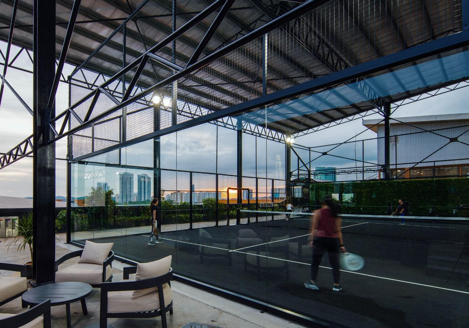 Indoor glass-walled padel court at Ascaro Padel & Social Club with players on court and a lounge seating area beside it