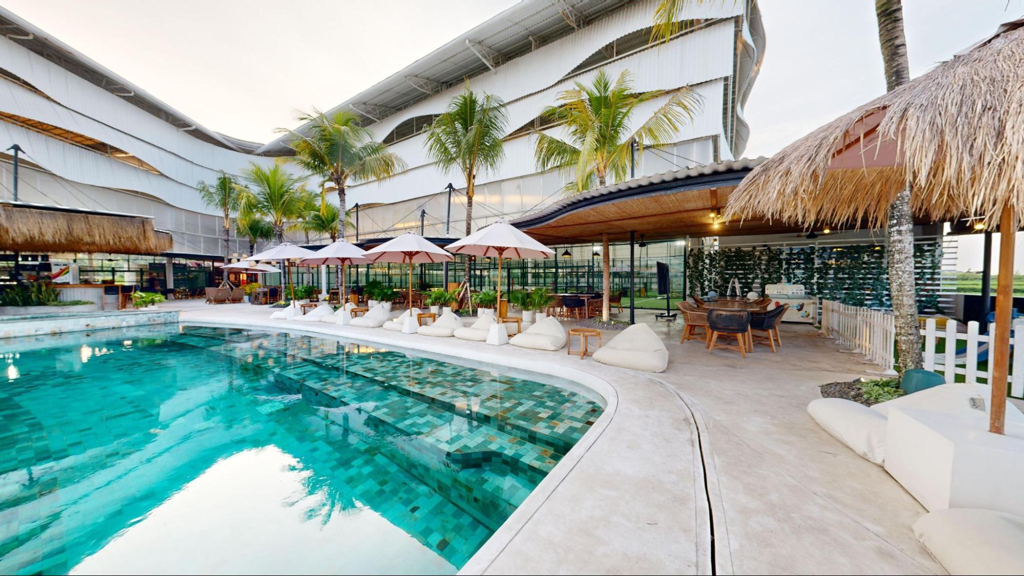 Outdoor pool and seating area with sun loungers and umbrellas, set in front of a modern multi-court padel facility surrounded by palm trees.