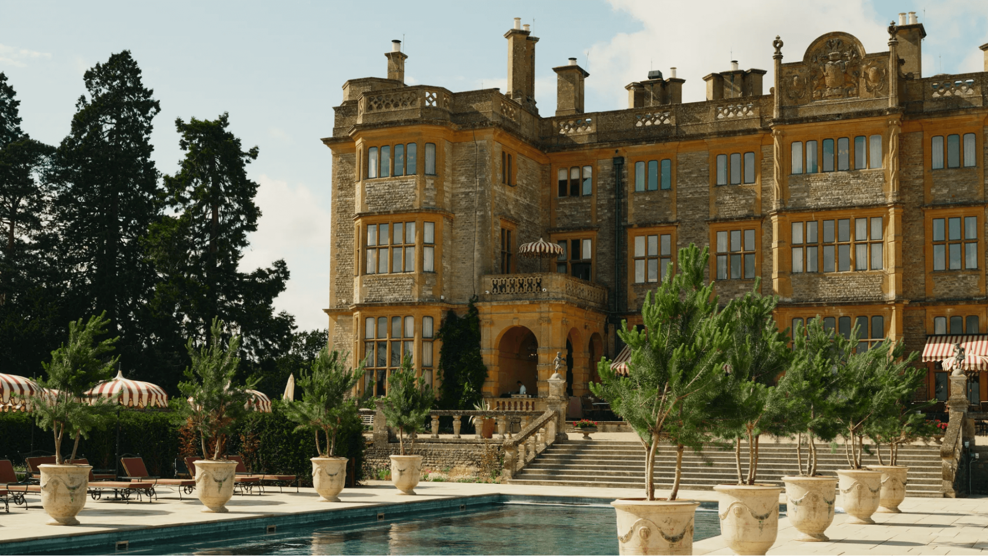 View of Estelle Manor in Oxfordshire, featuring a grand country house, outdoor pool, and surrounding trees in a luxury countryside setting.