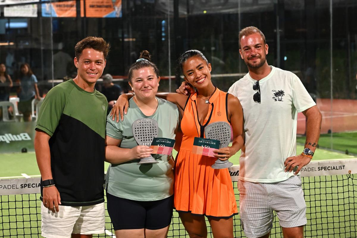 Four players at The Cage Padel Tribe standing in front of the net, smiling and holding prize envelopes after a match.