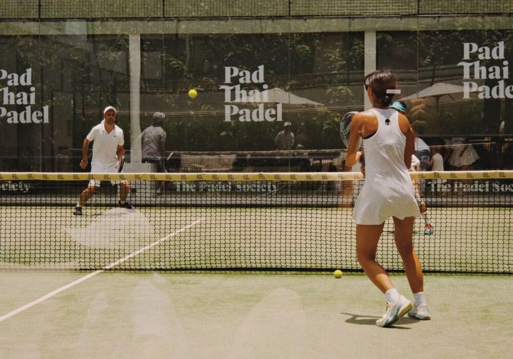 Two players rally on a sunny court at Pad Thai Padel in Bangkok, Thailand, surrounded by branded walls and shaded seating areas