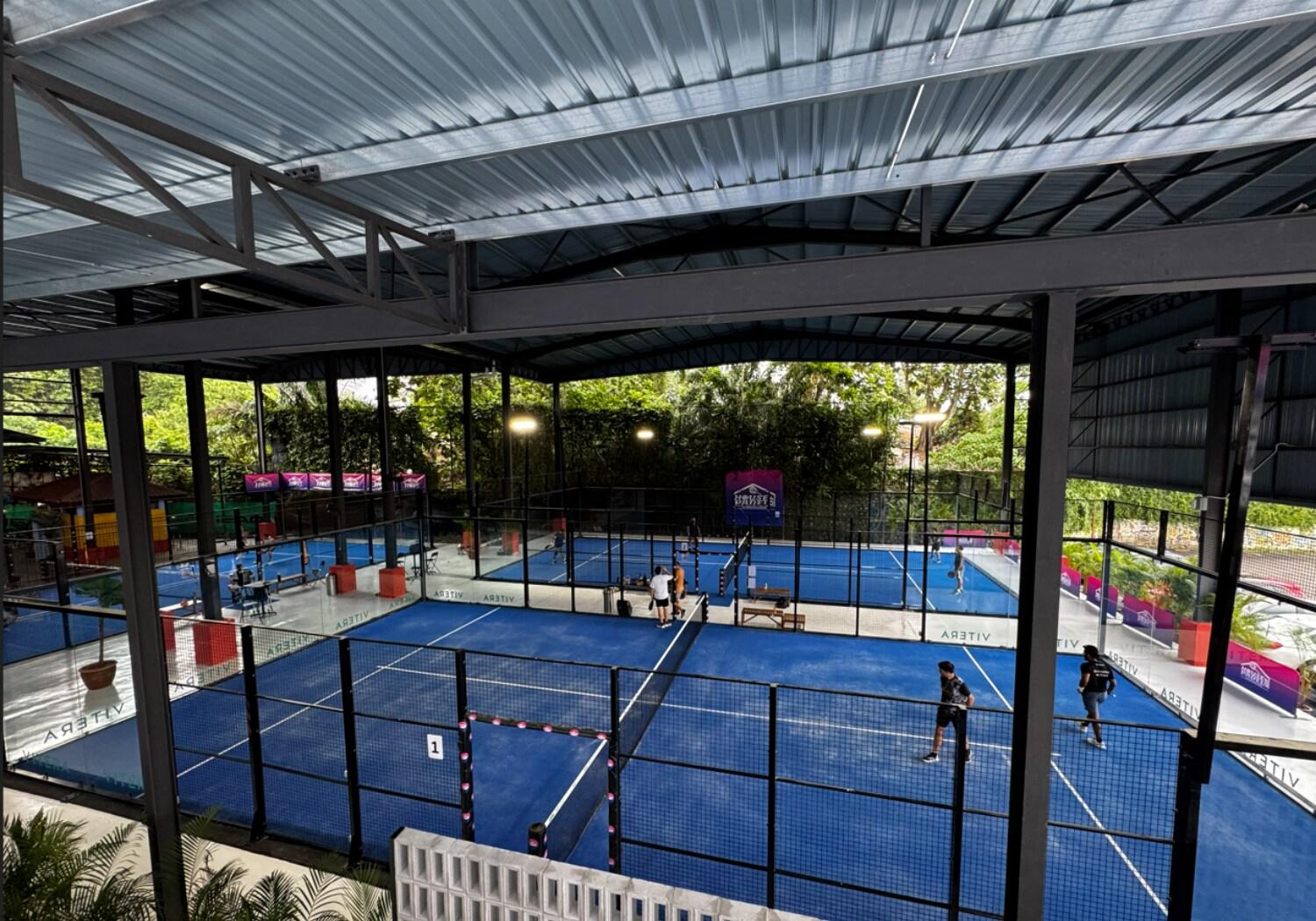 Multiple blue indoor padel courts at House of Padel with players training under a covered structure surrounded by greenery