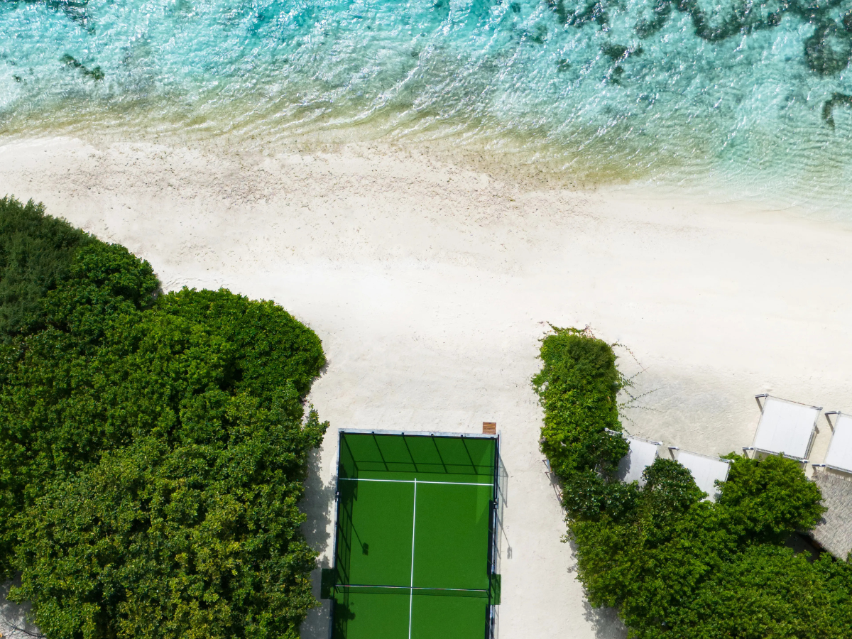 Aerial view of a scenic padel court between lush greenery and a turquoise ocean on a white-sand beach.