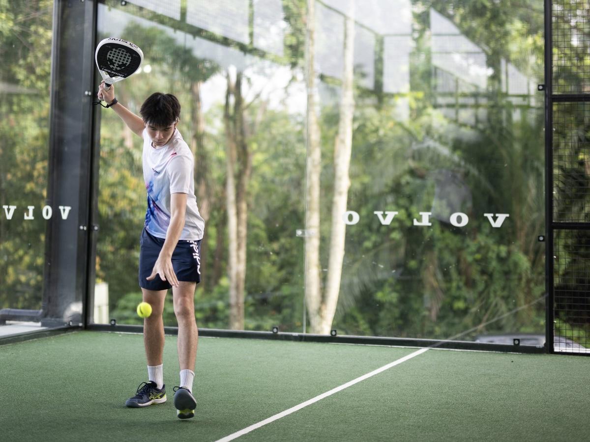 Male padel player on a green court bouncing the ball and preparing to serve with racket raised overhead.
