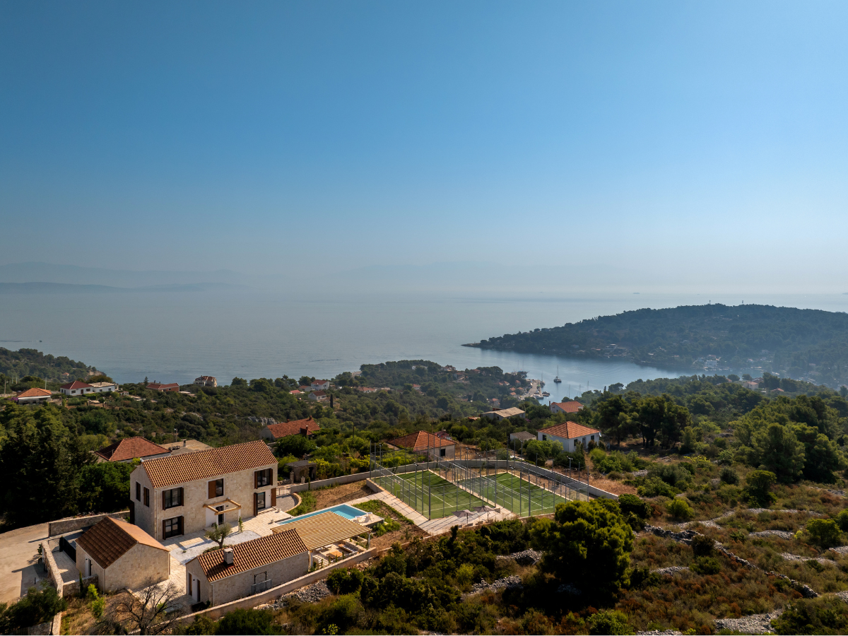 Hilltop padel court and villa overlooking the sea, with views of a coastal village, forested hills, and a calm blue horizon under a clear sky.