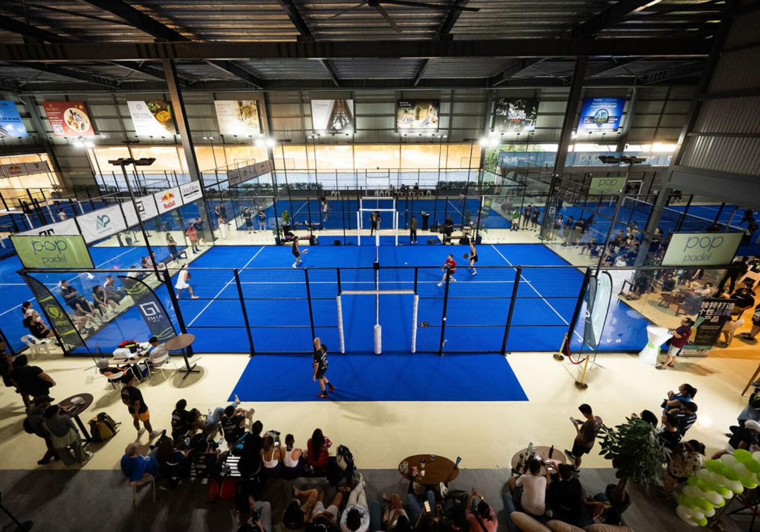 Large indoor padel venue at Pop Padel with multiple blue courts, players competing, and spectators watching from seating areas