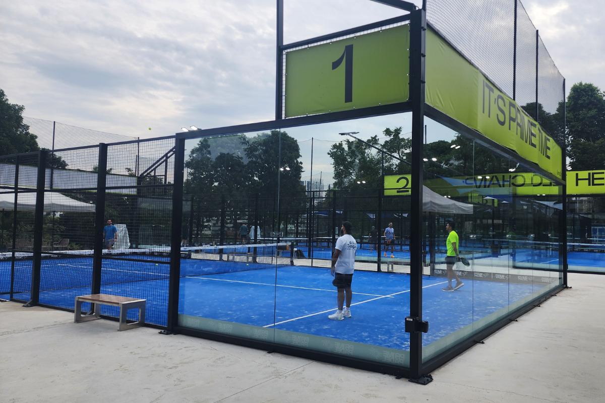A blue glass-walled padel court at Prime Padel Dempsey, with players on court and trees in the background.
