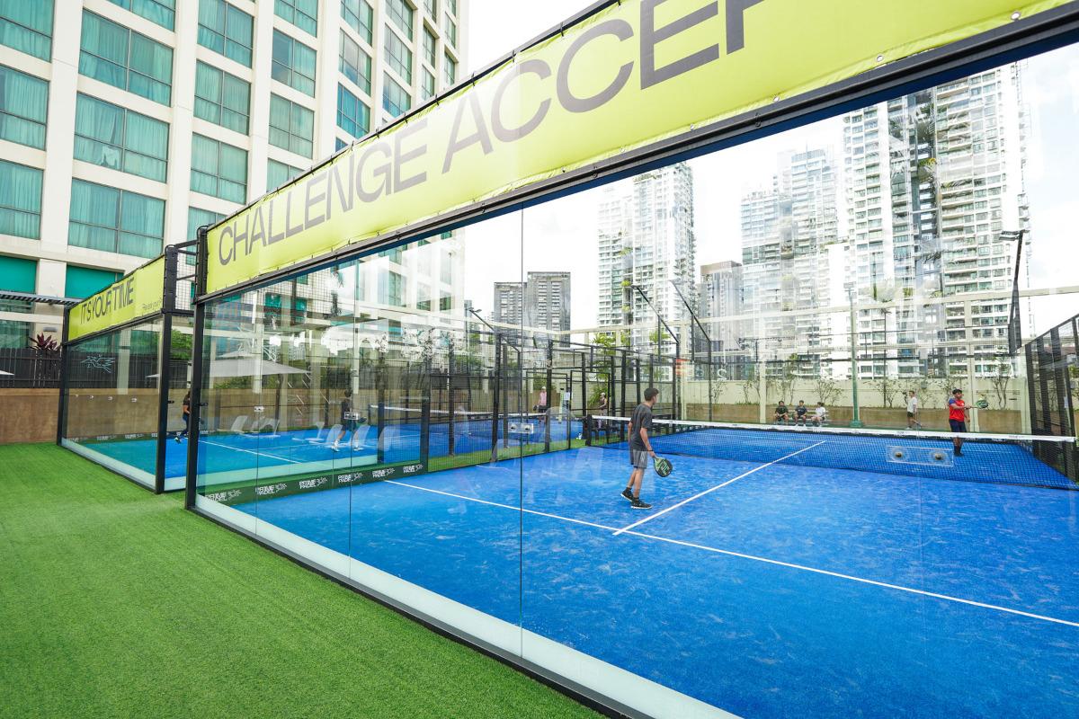 A blue glass-walled padel court at Prime Padel Grand Copthorne, set against tall city buildings with players on court.