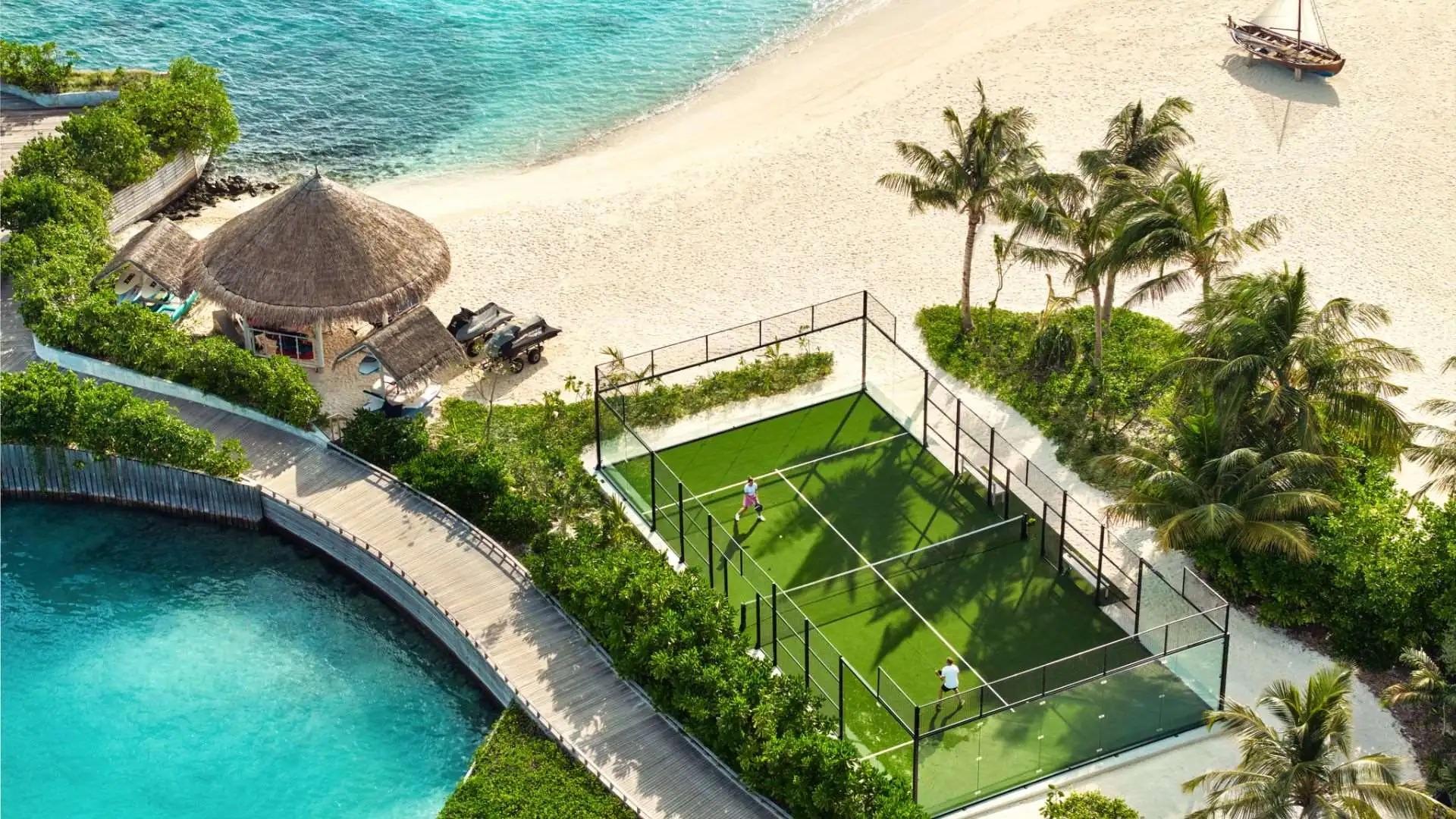 A beachfront padel court at The Nautilus Maldives, set on white sand and surrounded by turquoise ocean waters and lush tropical greenery.